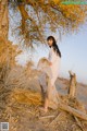 A woman in a white dress and hat standing next to a tree.